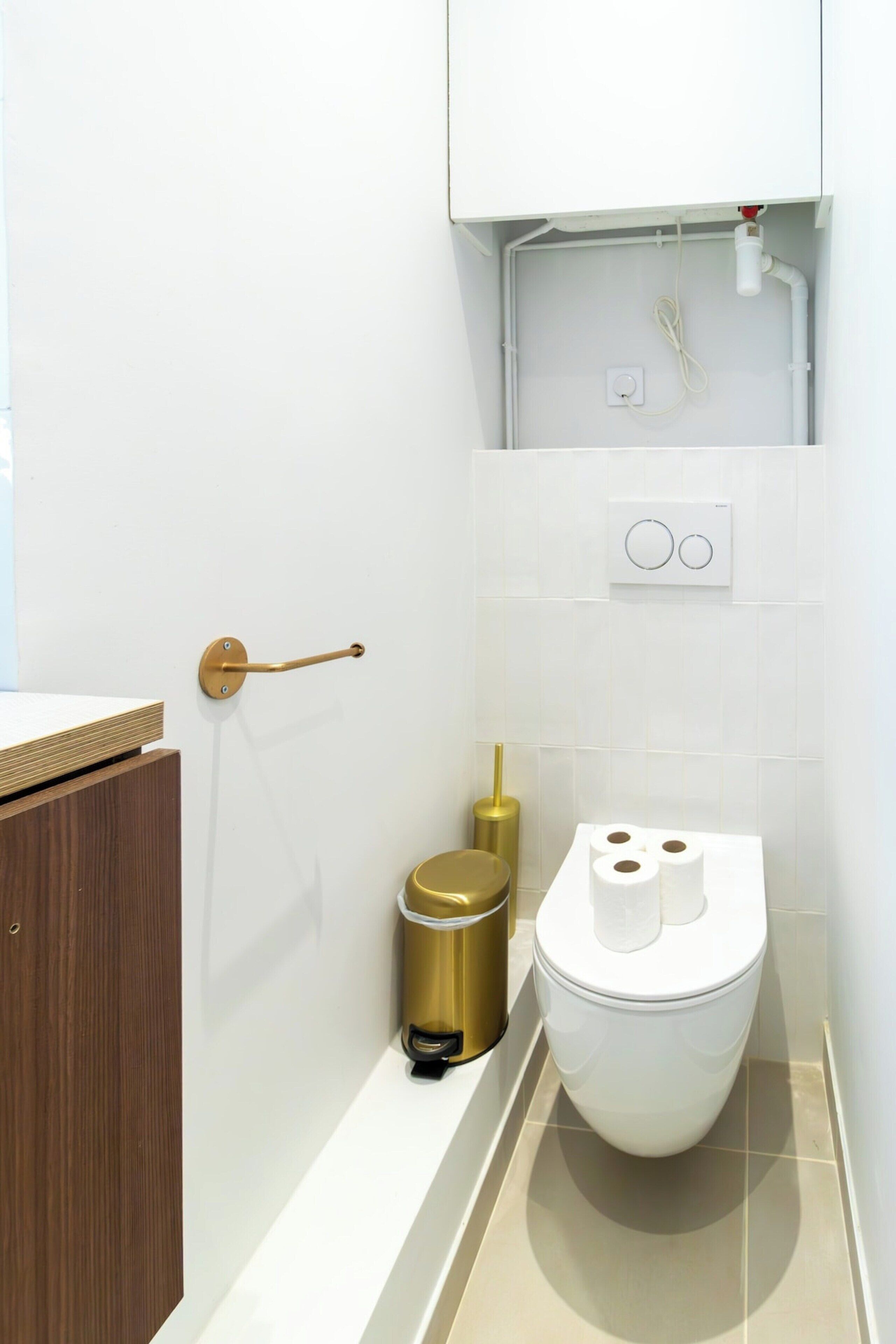 A modern bathroom with a toilet, a gold trash bin, a toilet paper holder, and a wooden cabinet.