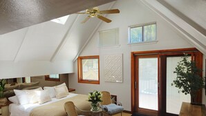Acogedor dormitorio en ático con cama, dos sillas, una mesa de madera, ventilador de techo y un gran ventanal con persianas.