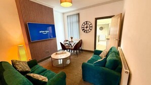 Ein Wohnzimmer mit grünen Sofas, einem runden Couchtisch, einem an der Wand montierten Fernseher, einem Esstisch mit Stühlen und einer Wanduhr.