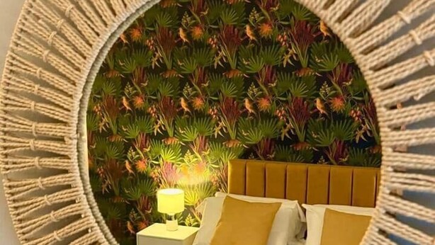 Ein runder Spiegel mit Makramee-Rahmen, der ein Bett mit Kopfteil, Kissen und einer Nachttischlampe reflektiert.