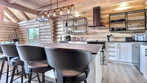 A modern log cabin interior with a central island dining table, bar stools, a kitchen with stainless steel appliances, and a hanging light fixture. - Spa, Game Room, Absolute Comfort | Le Commandant (Saint-Philémon)