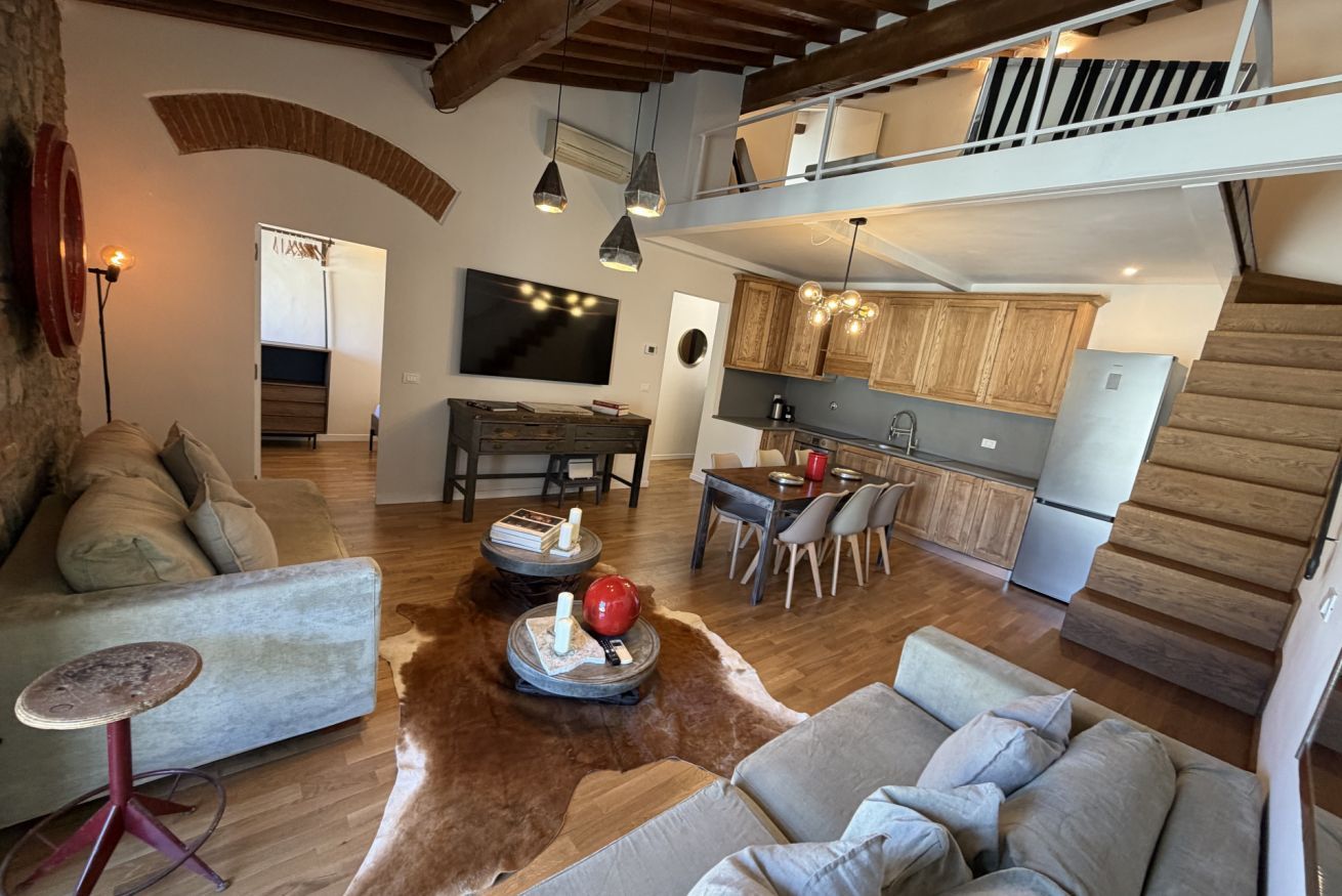 A spacious living area with a sofa, a coffee table, and a dining table. A kitchen is visible in the background with wooden cabinets and a refrigerator.