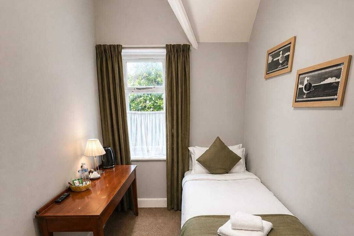 Quarto pequeno e bem organizado com cama, escrivaninha, um banco e janela com cortinas.