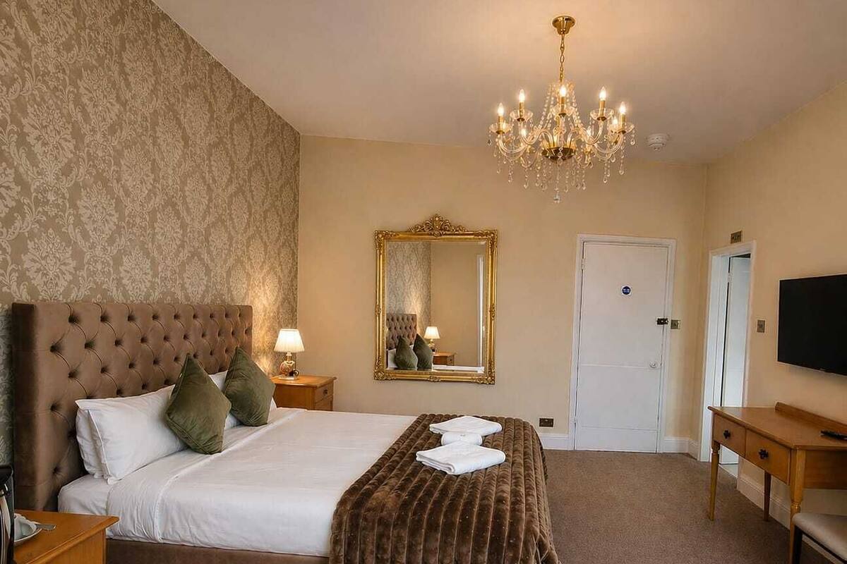 Quarto de hotel com uma cama grande, um candelabro, um espelho, uma televisão e uma mesa de cabeceira de madeira.