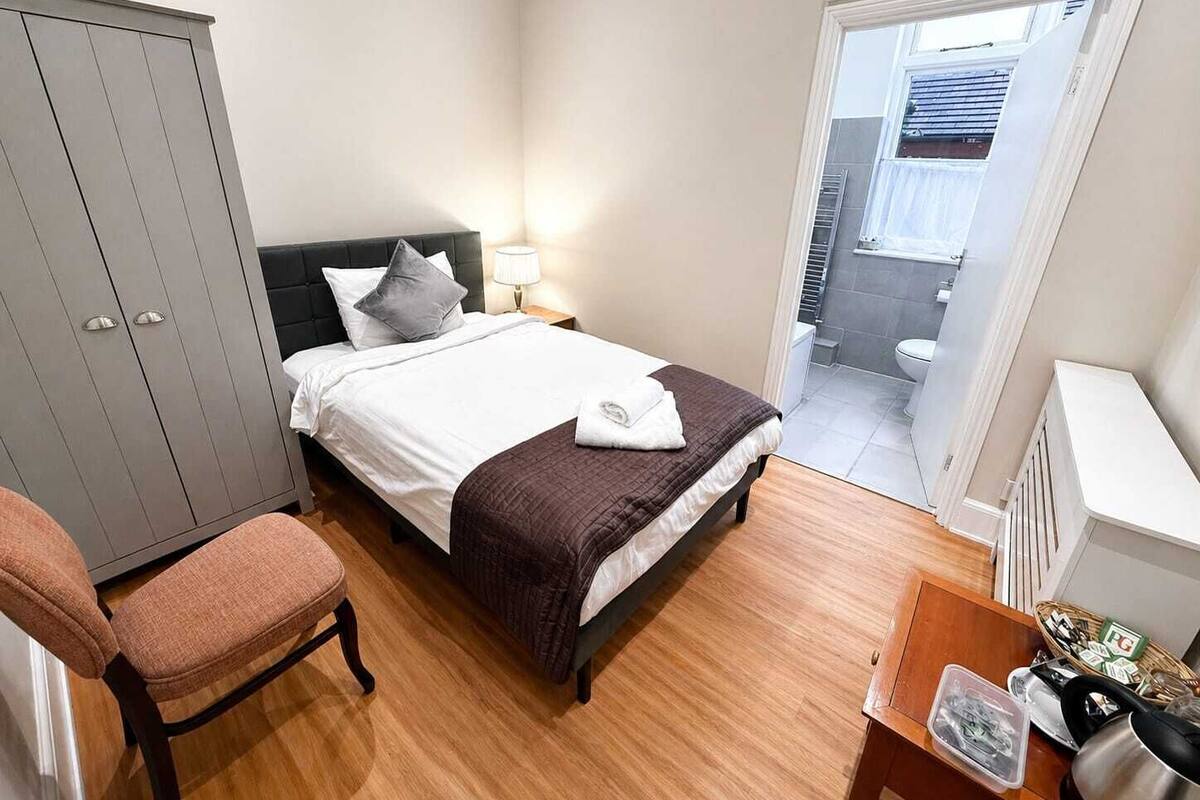 Quarto com cama, cadeira, guarda-roupa, mesa de cabeceira com abajur e vista para o banheiro.