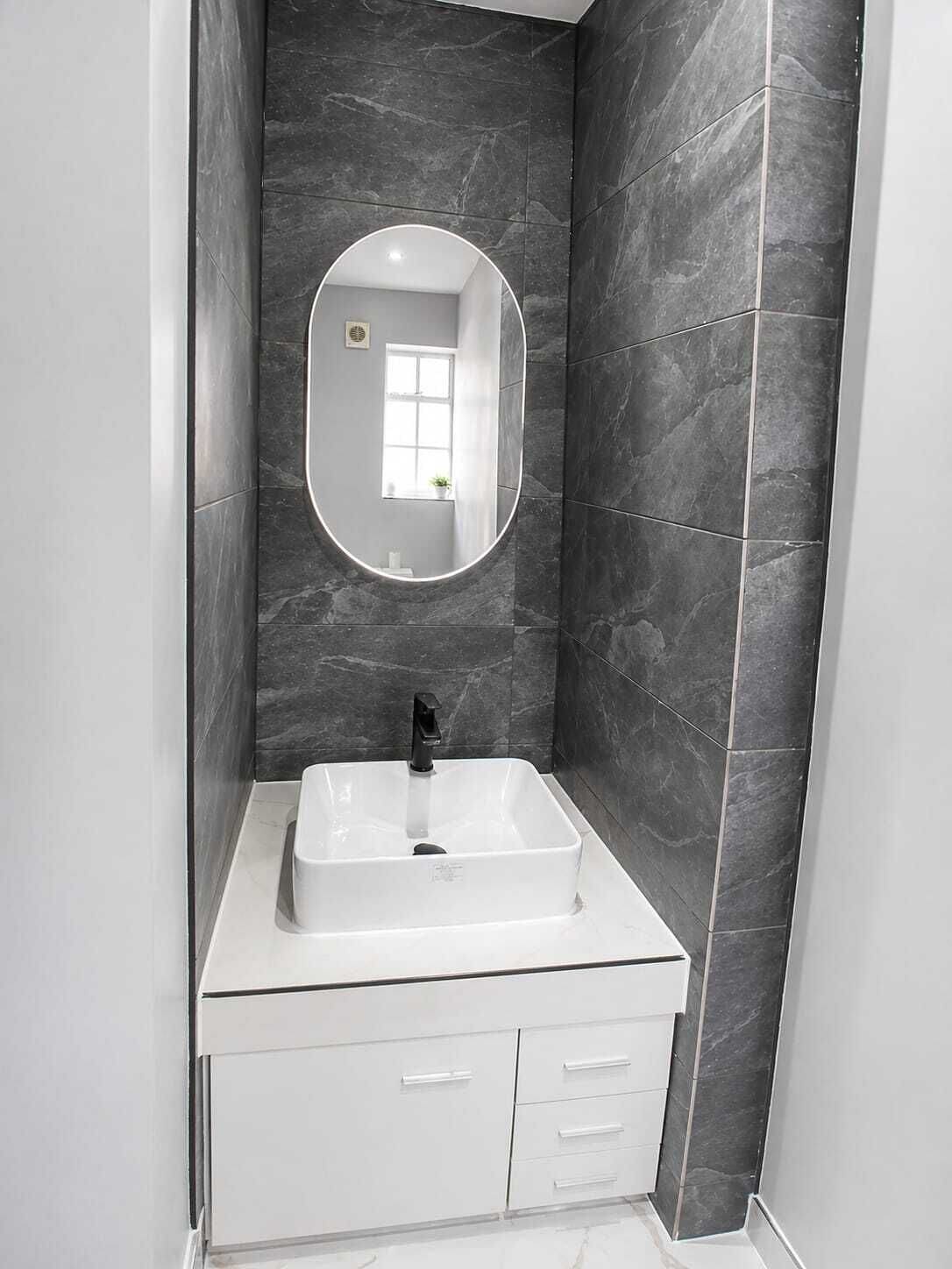 Un baño con tocador blanco, grifo negro, espejo grande y paredes de mármol oscuro.
