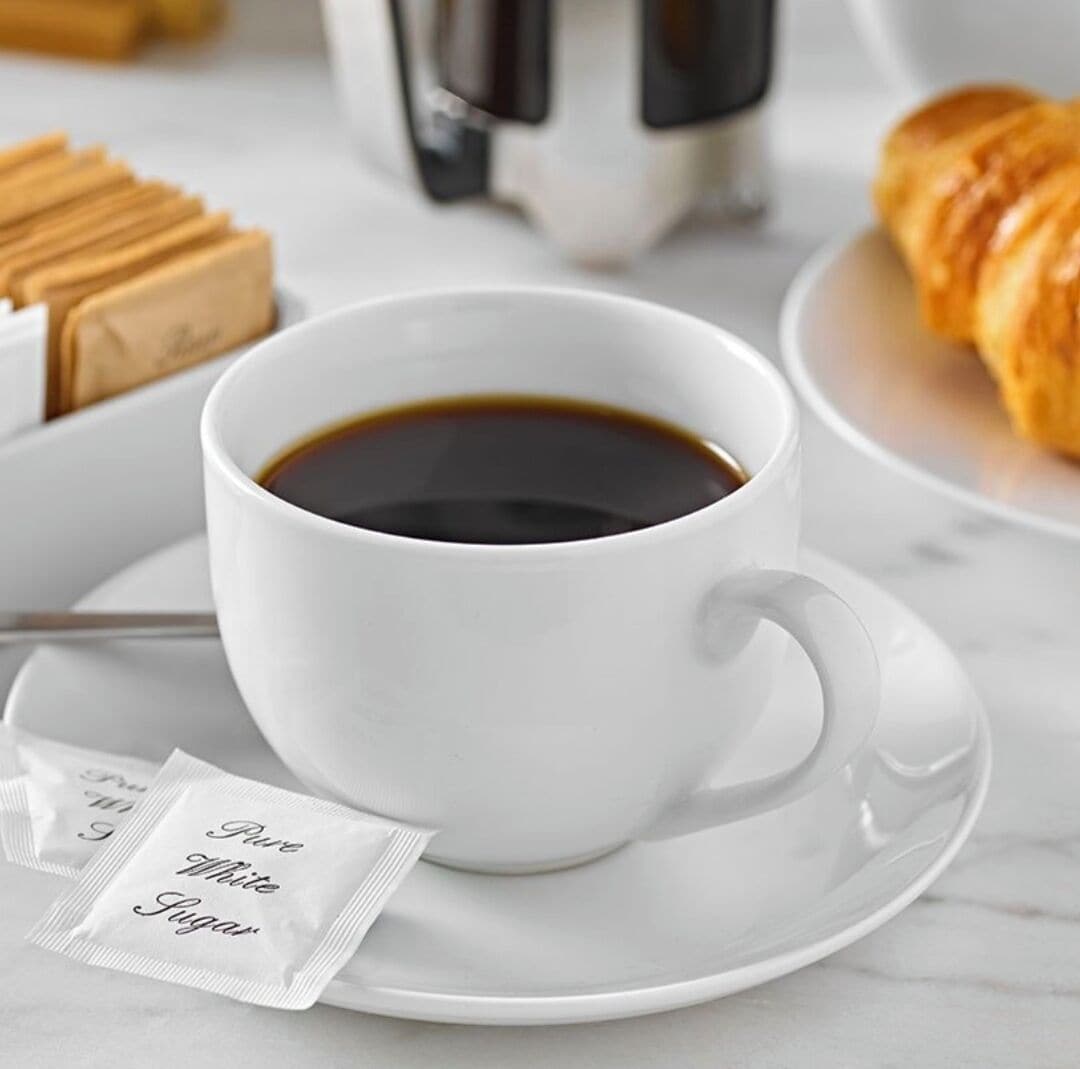 A white cup of coffee with a saucer, a packet of Pure White Sugar, and a croissant on a marble surface.