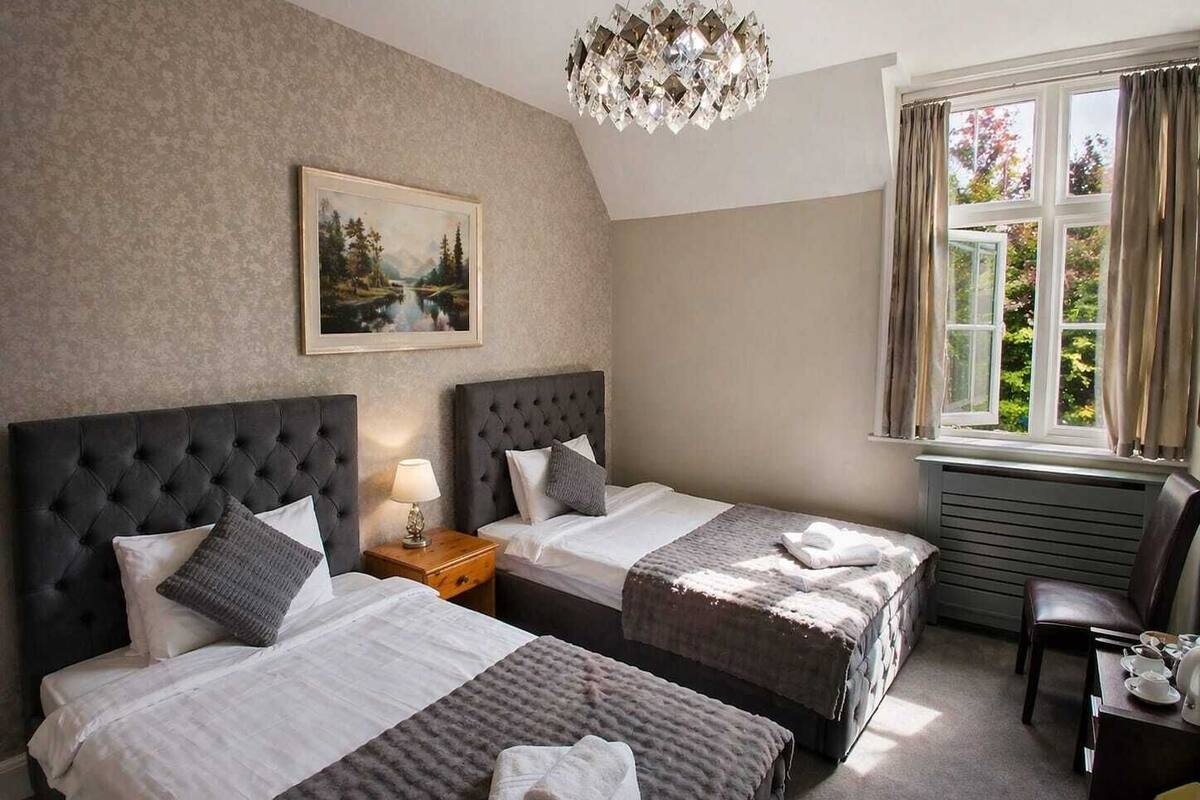 Quarto de hotel com duas camas, um quadro na parede, um lustre e uma janela com cortinas.