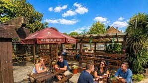 An outdoor patio with wooden tables and benches, a red umbrella labeled 'Old Speckled Hen,' and a brick building in the background. - Tree Hotel (Oxford)