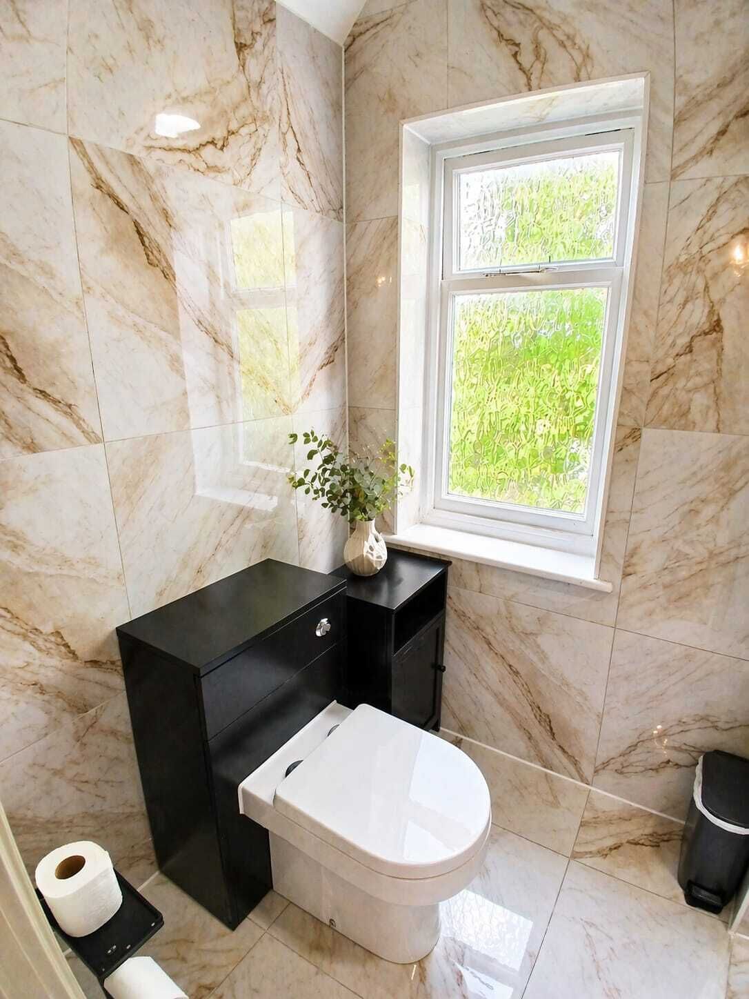 A bathroom with marble walls, a black vanity, a toilet, and a window with a view of greenery.