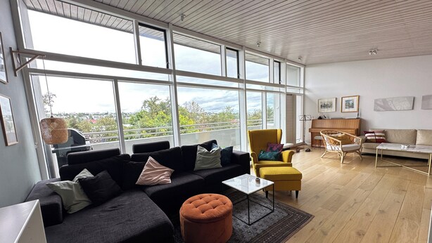 A spacious living room with a large window, a black sofa, a yellow armchair, and a wooden floor. - Modern and family friendly house in Fossvogur in Reykjavik (Reykjavík)