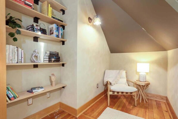 A cozy attic room with wooden flooring, a white chair, a small table with a lamp, and bookshelves filled with books.
