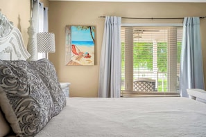 Un dormitorio con cama, un cuadro de una playa, una ventana con persianas y vistas a zonas verdes.