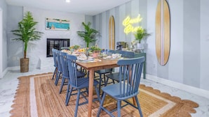 A dining area with a wooden table, blue chairs, and a rug. There are two surfboards on the wall and a neon sign that says "Beach Breeze."