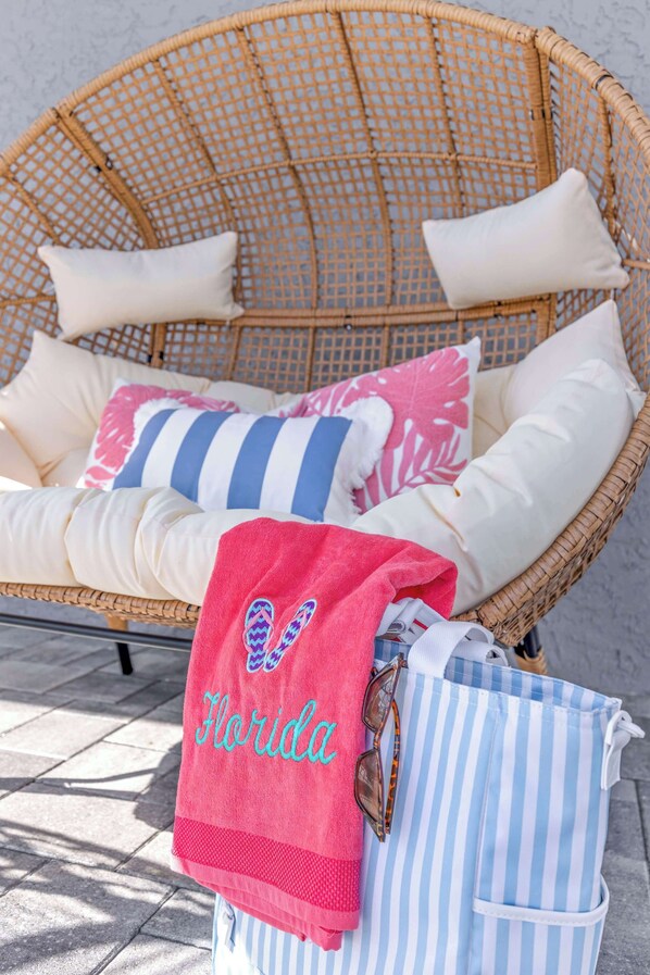 Uma cadeira de vime com almofadas brancas e travesseiros listrados, uma toalha rosa com a palavra 'Florida' bordada e uma bolsa listrada.