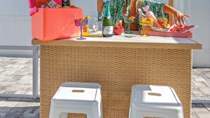 A beach bar setup with a thatched roof, wicker table, and white stools.