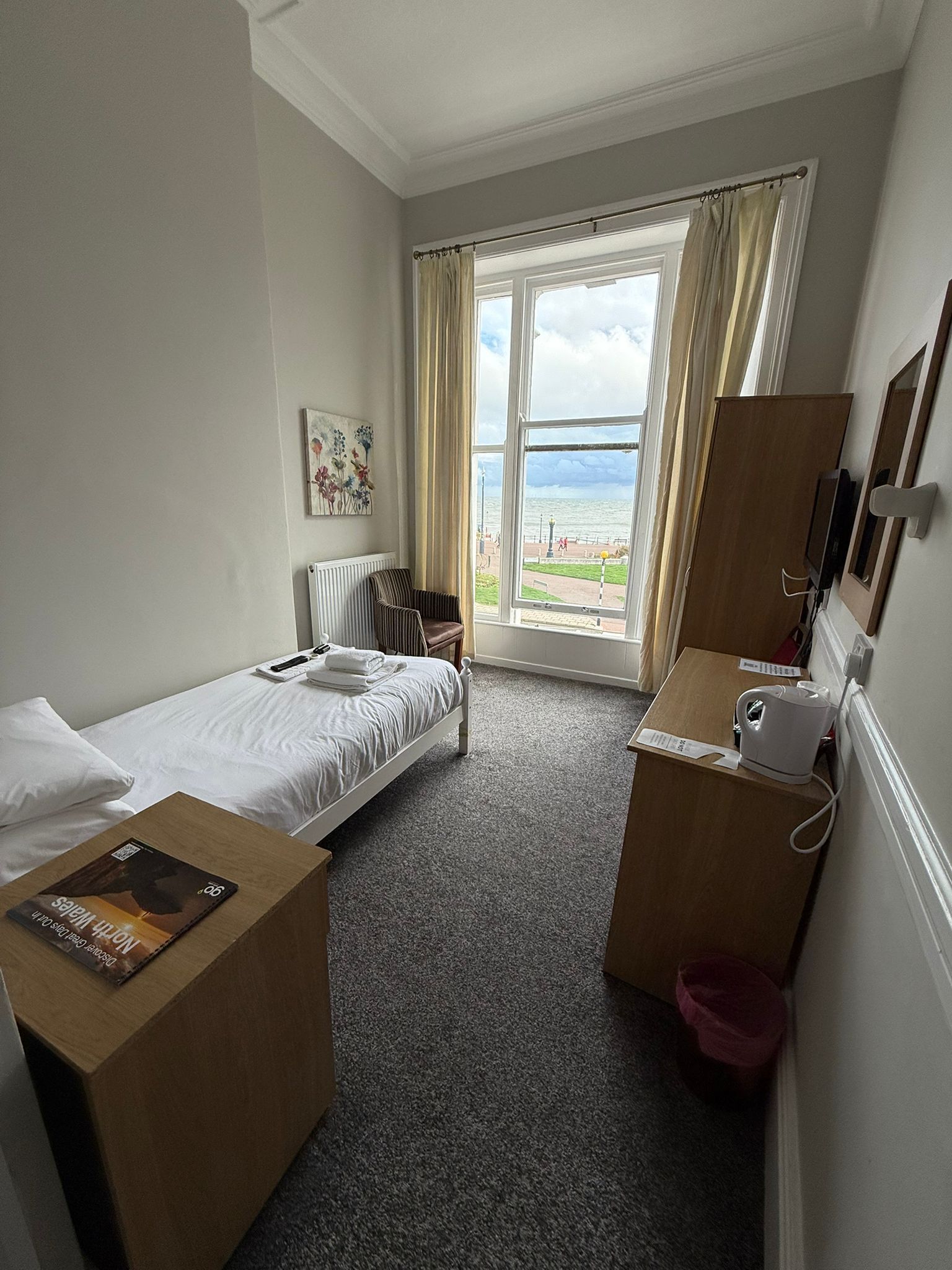 A hotel room with a bed, a small desk, a chair, a radiator, a window with curtains, a painting on the wall, and a view of the sea.