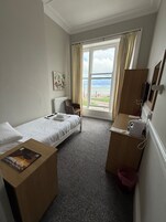 A hotel room with a bed, a small desk, a chair, a radiator, a window with curtains, a painting on the wall, and a view of the sea.