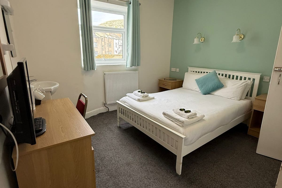 A hotel room with a bed, a desk with a computer, a window with curtains, and a radiator.