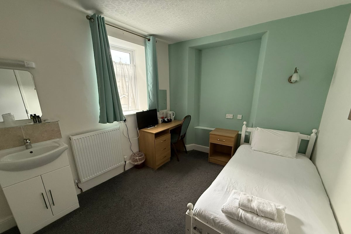 A bedroom with a bed, a desk, a chair, a small bedside table, a radiator, a window with curtains, a mirror, and a sink.