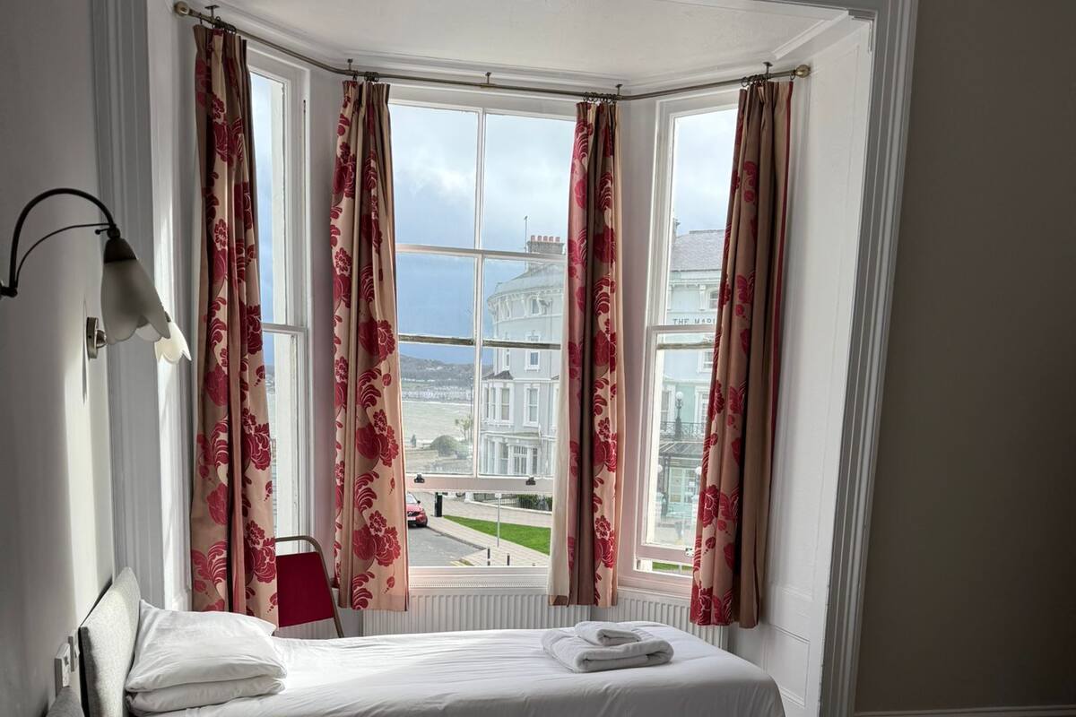 A hotel room with two beds, a view of a building through large windows, and a lamp on the left side.