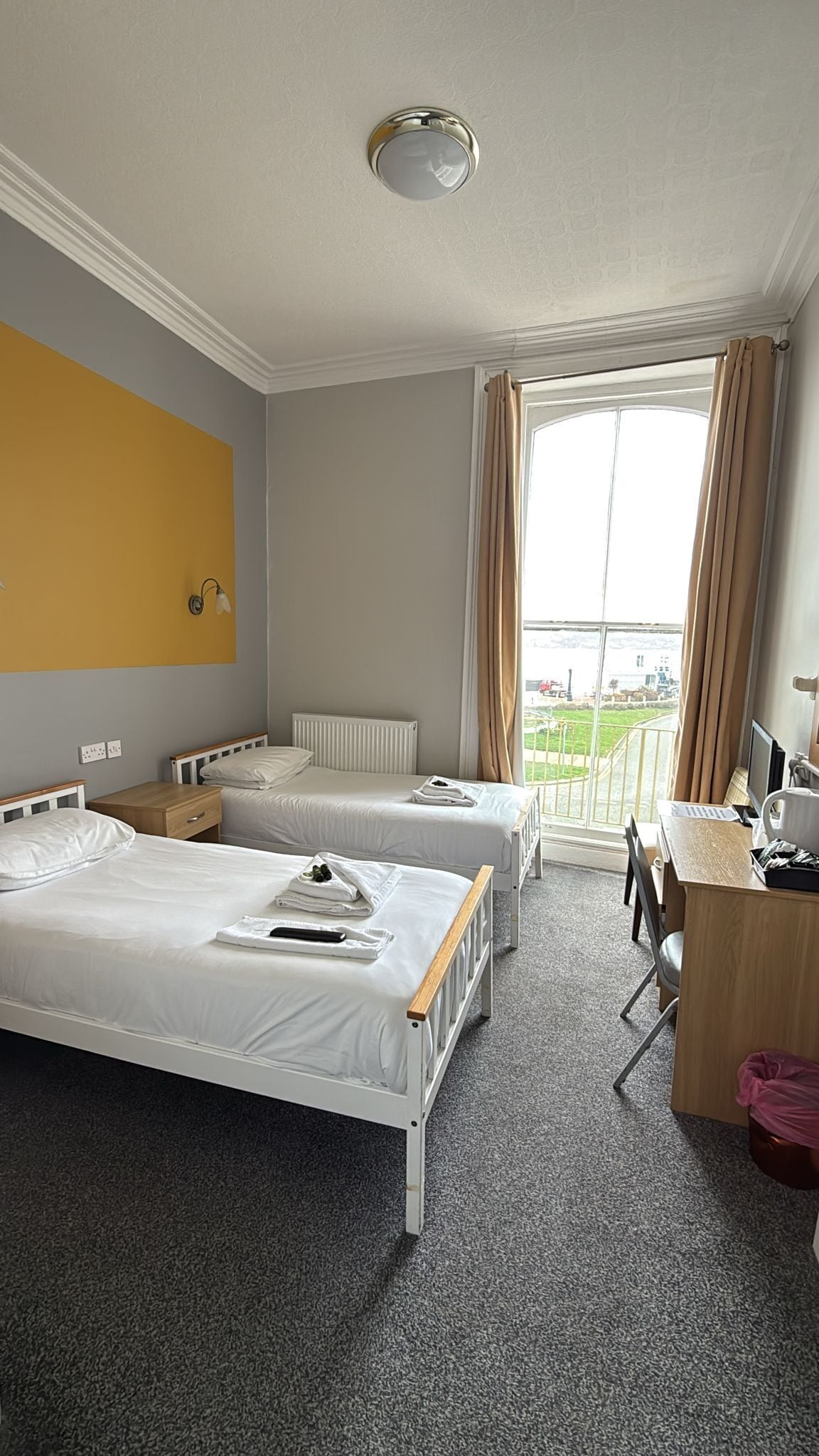 A hotel room with two single beds, a desk with a computer, a window with curtains, and a view of the outside.