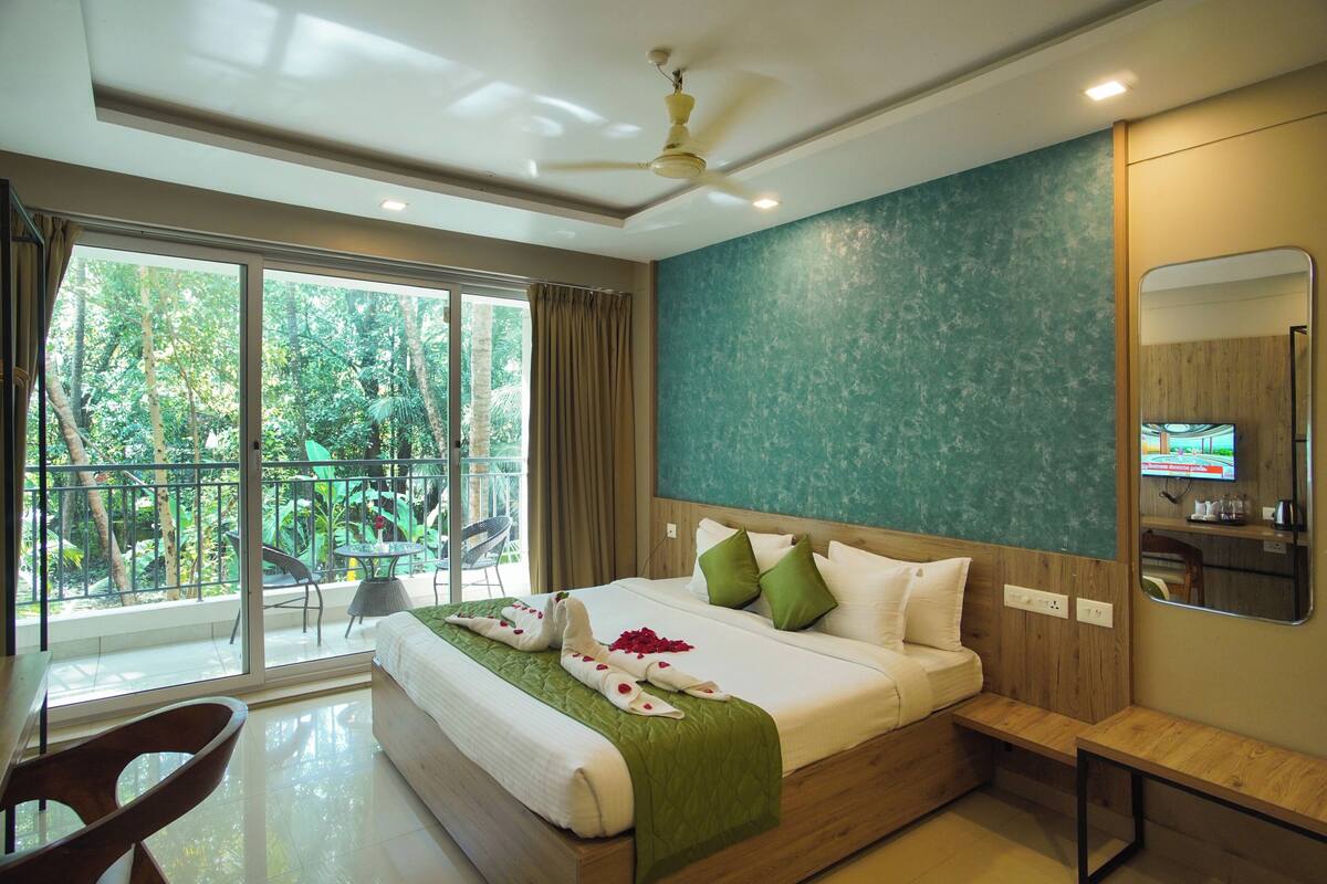 A hotel room with a large bed, a ceiling fan, a TV, and a balcony with a view of trees.