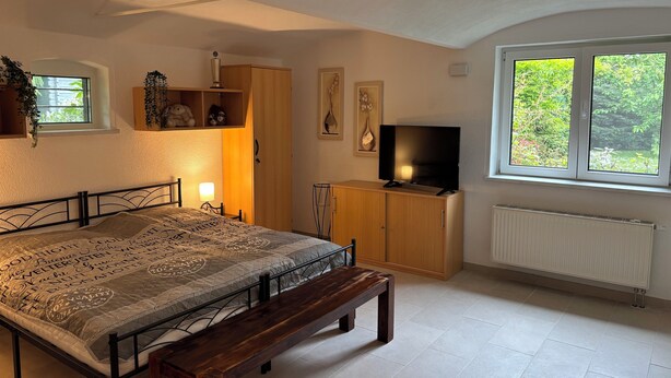 A bedroom with a bed, a bench, a TV, a radiator, and a window with a view of greenery. - Cozy vacation apartment at Ammelshain Castle (Naunhof)