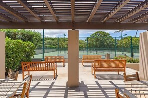 A covered outdoor seating area with wooden benches and tables, overlooking a tennis court. - Fully Remodeled Oceanfront Condo MAKENA SURF RESORT, #F-206 (Makena, Maui)