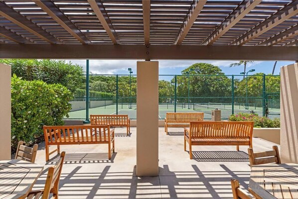 A covered outdoor seating area with wooden benches and tables, overlooking a tennis court.