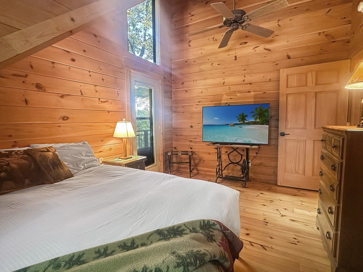 A wooden cabin bedroom with a bed, a television on a stand, a ceiling fan, and a dresser.
