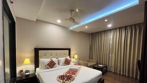 A hotel room with a large bed, a ceiling fan, a bedside table with a lamp, and a view of the outside through a window. - Hotel The Westinn (Varanasi)