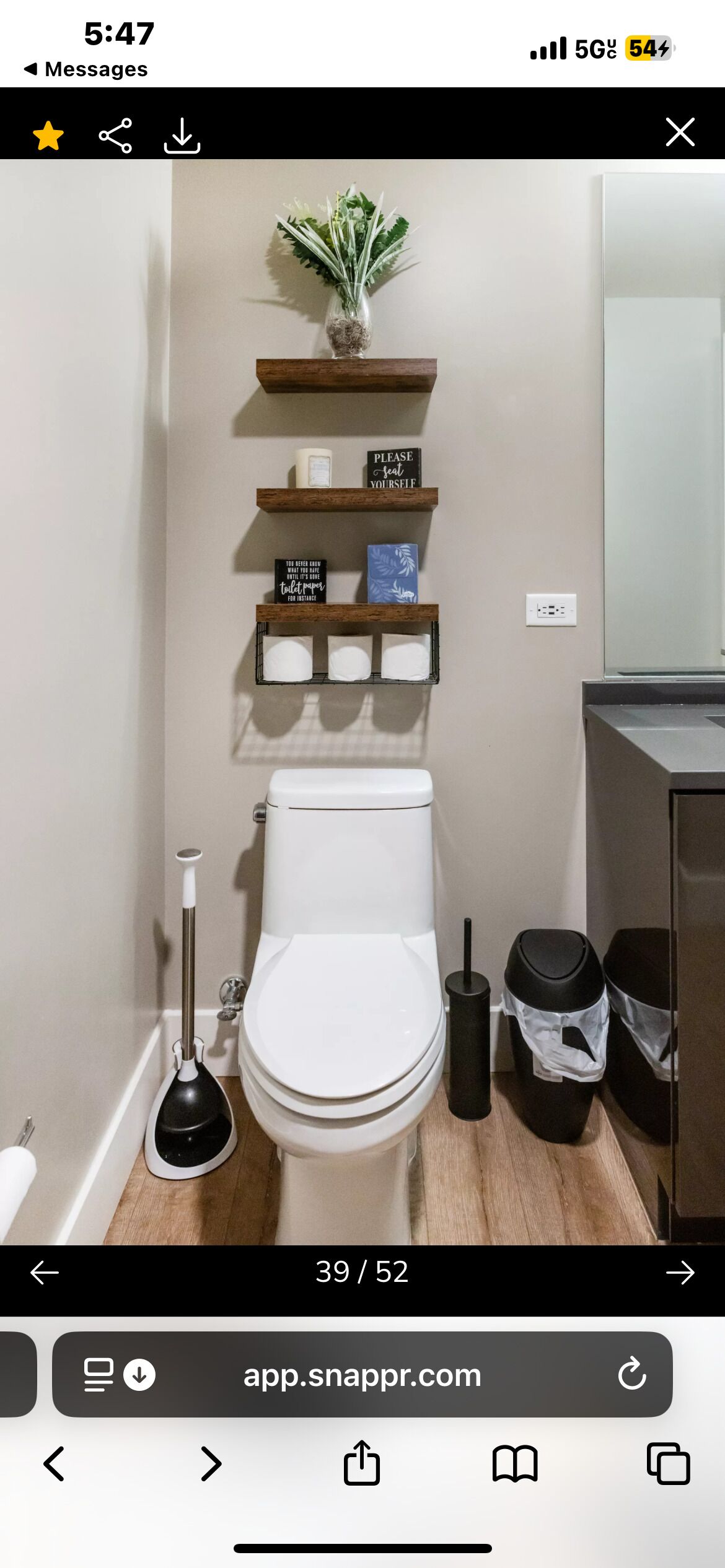 A modern bathroom with a toilet, a plunger, a trash bin, and a shelf with toiletries.