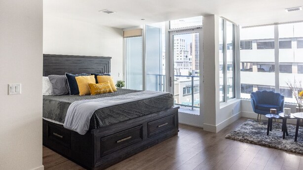 Une chambre moderne avec un grand lit, une vue sur la ville grâce à de grandes fenêtres et un balcon agrémenté d’une chaise et d’une table.