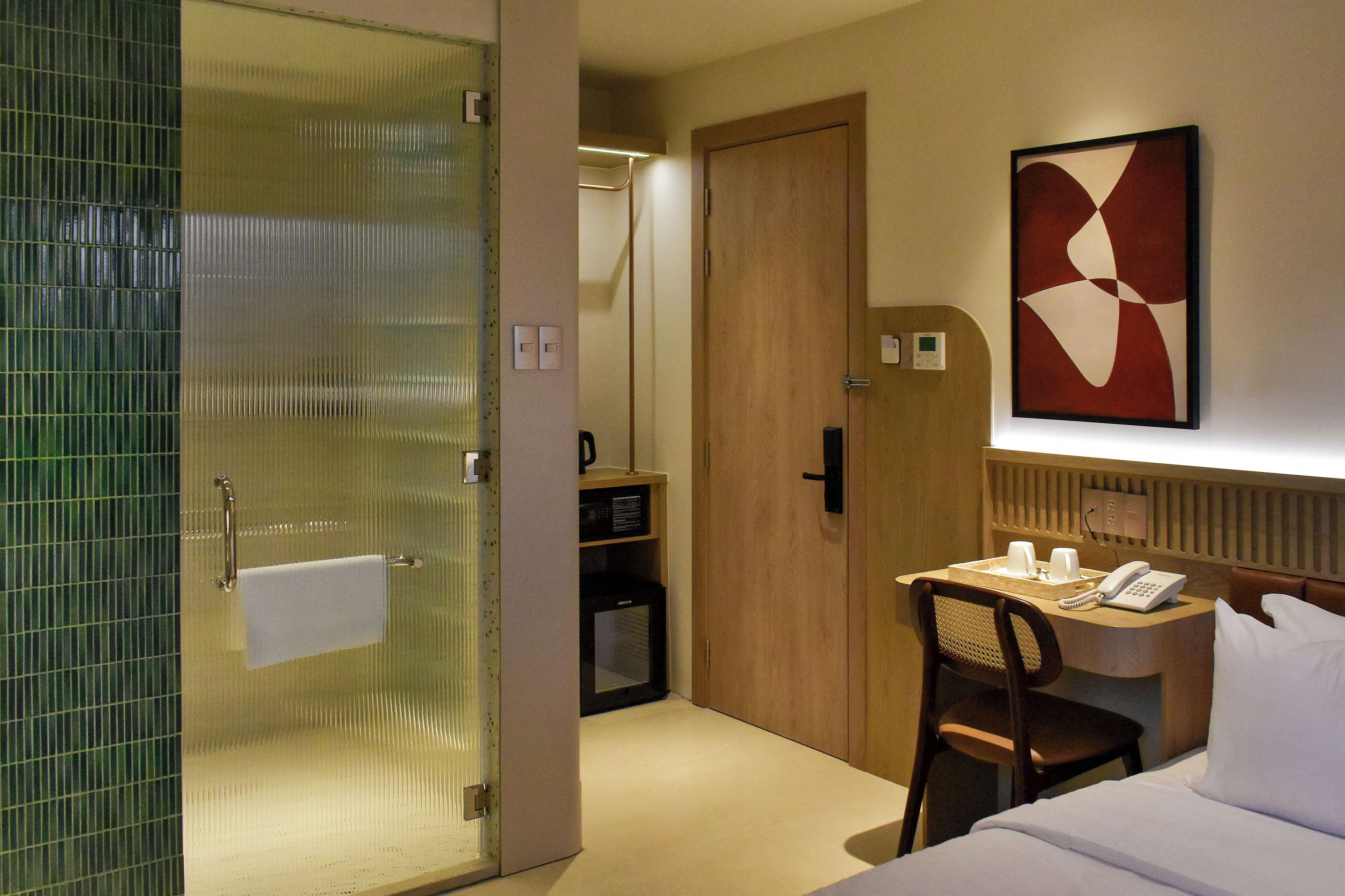 Een hotelkamer met een douchecabine van glas, een houten deur, een bureau met telefoon en een bed met wit beddengoed.