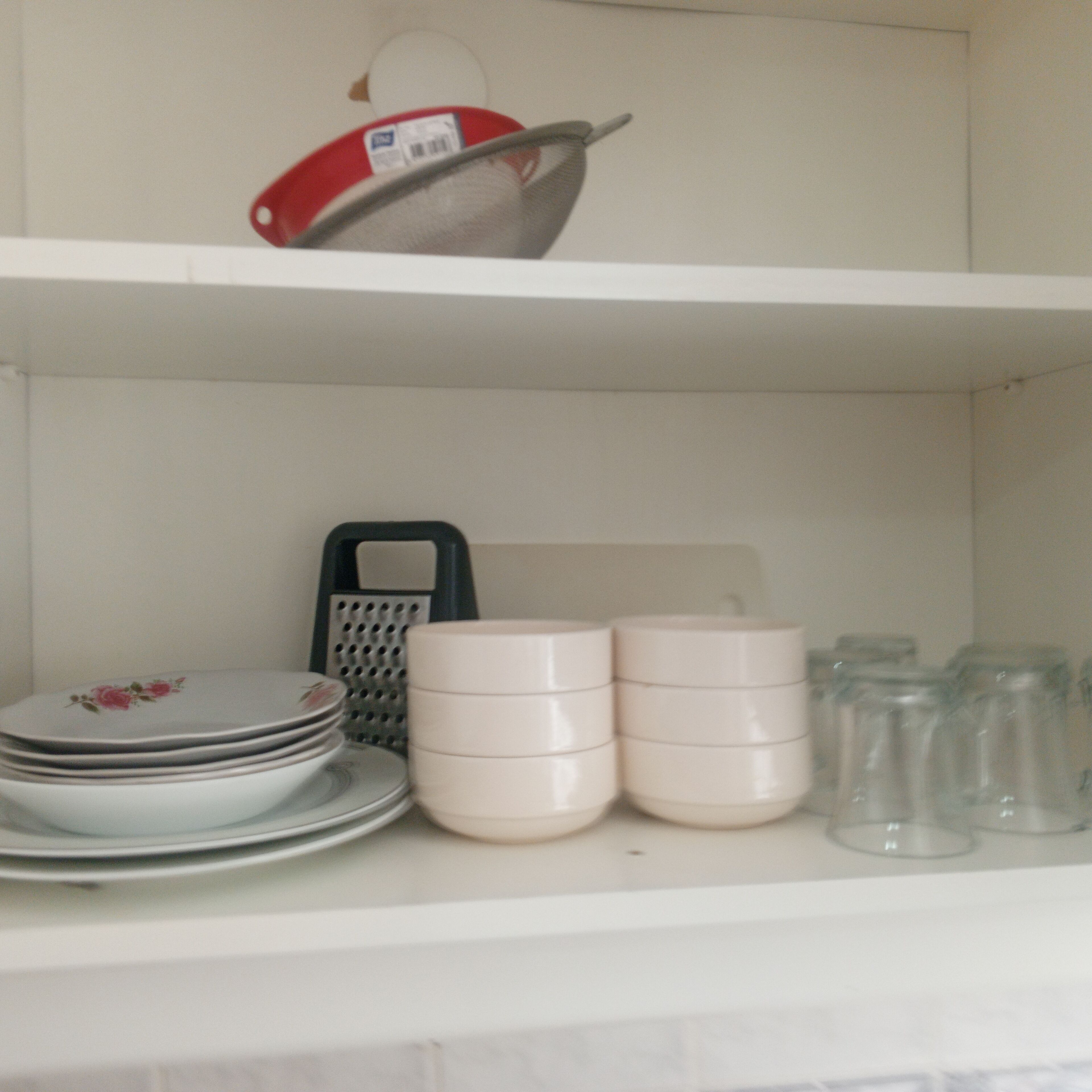 Una estantería en la cocina con platos apilados, un colador, un rallador y varios frascos de vidrio.