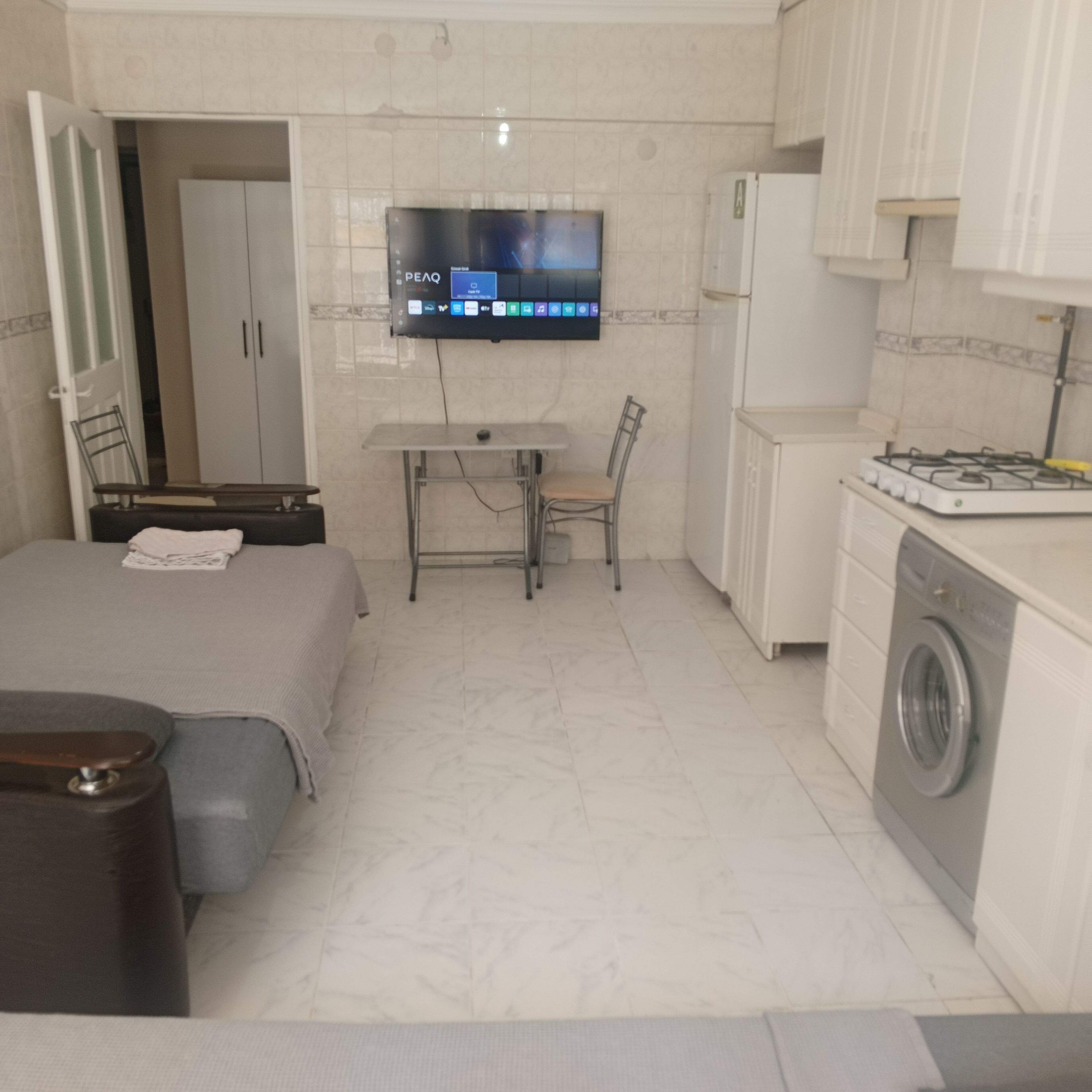 A compact hotel room with a bed, a desk, a chair, a washing machine, and a television mounted on the wall.