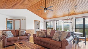 A spacious living room with two brown leather sofas, a wooden coffee table, and a ceiling fan. Large windows offer a view of the outdoors. - Everview 3 BR 3 BATH Spectacular Mountain Views Firepit, Wi-Fi, Free Pool Access (Townsend)
