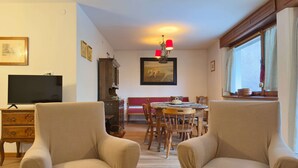 A living room with a television, a dining table with chairs, a chandelier, and a window with curtains. - Ca' Del Molin, spacious apartment with view (Cortina d'Ampezzo)