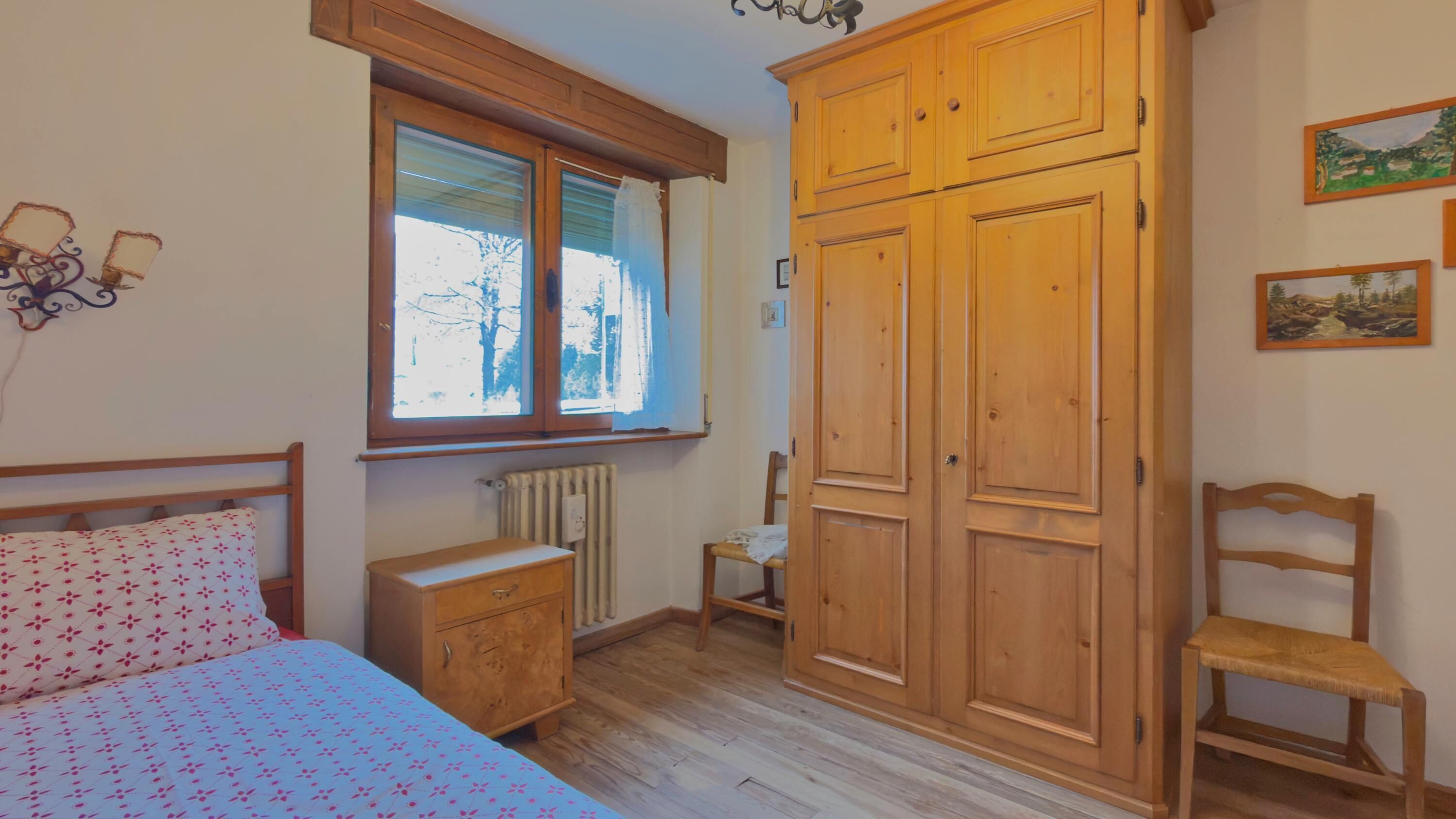 A bedroom with a bed, wooden wardrobe, a chair, a radiator, and a window with blinds.
