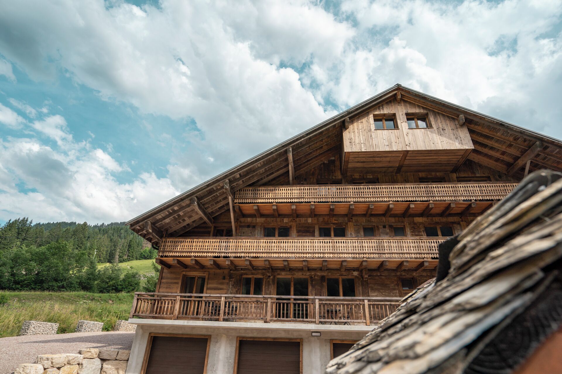 A wooden chalet with a balcony, a garage, and a mountainous backdrop.
