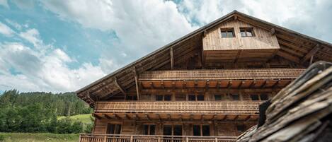 A wooden chalet with a balcony, a garage, and a mountainous backdrop.