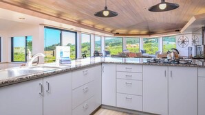 Una cucina moderna con mobili bianchi, piani di lavoro in granito e un'isola grande. Si vedono due luci a sospensione, un fornello e un lavello. La cucina offre vista su un paesaggio suggestivo attraverso ampie finestre.