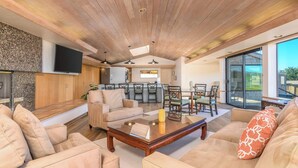 Un'ampia zona giorno con soffitto in legno, una TV a schermo piatto fissata al muro, un'area pranzo con tavolo e sedie in vetro, e un'ampia porta scorrevole in vetro che dà su un patio esterno.