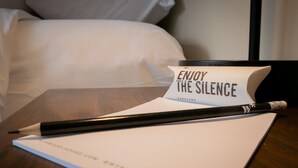 Notitieboekje met pen op een houten tafel, een bordje met de tekst 'ENJOY THE SILENCE' en een bed met witte lakens op de achtergrond.