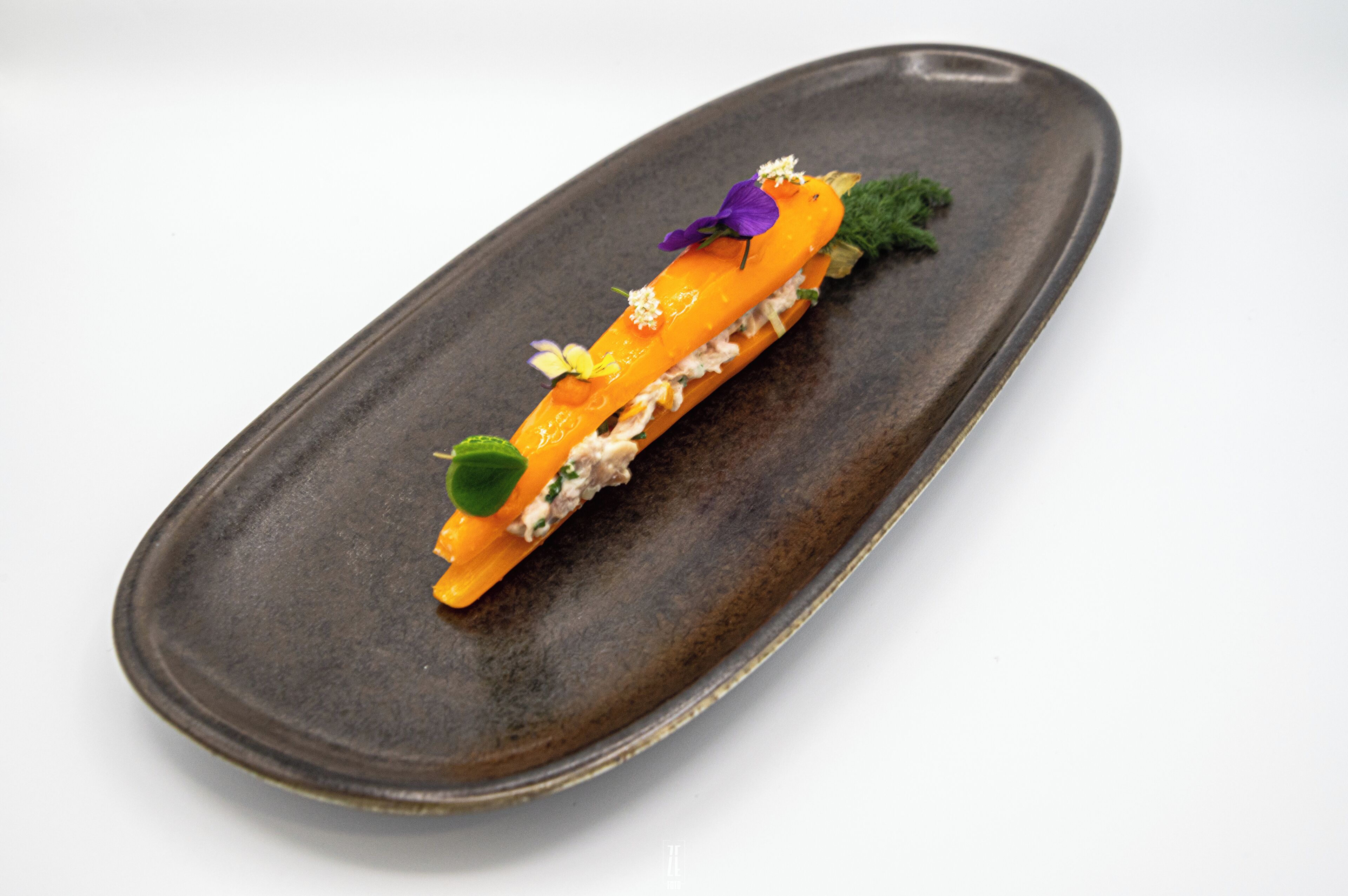 Une assiette en céramique de couleur sombre, sur laquelle est présentée une seule portion de nourriture, peut-être un poisson ou une viande, agrémentée d’herbes et d’une fleur violette.