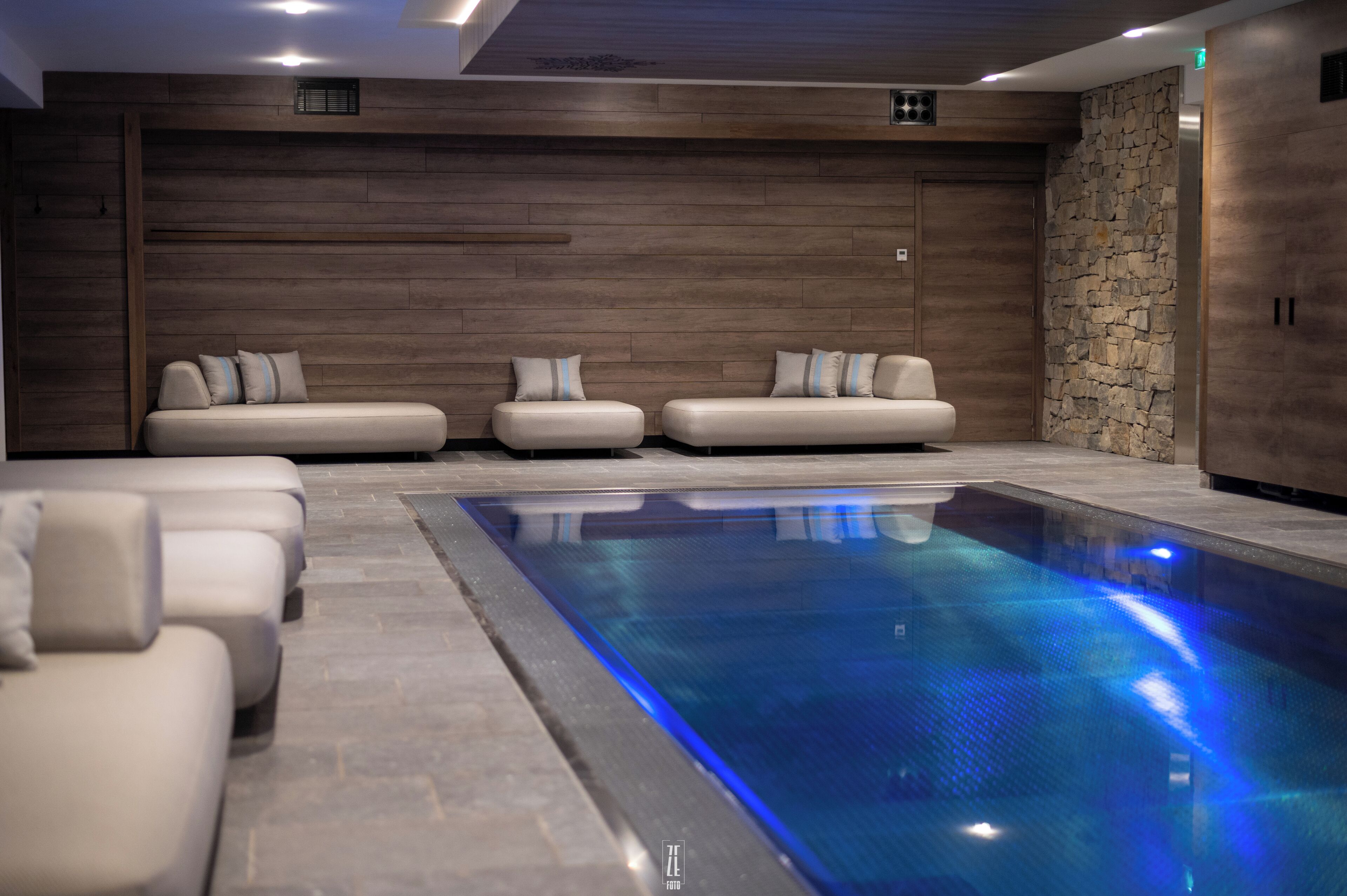 Ein moderner Poolbereich im Innenbereich mit einem langen, rechteckigen Pool, zwei Sofas mit Kissen und einer Steinwand.