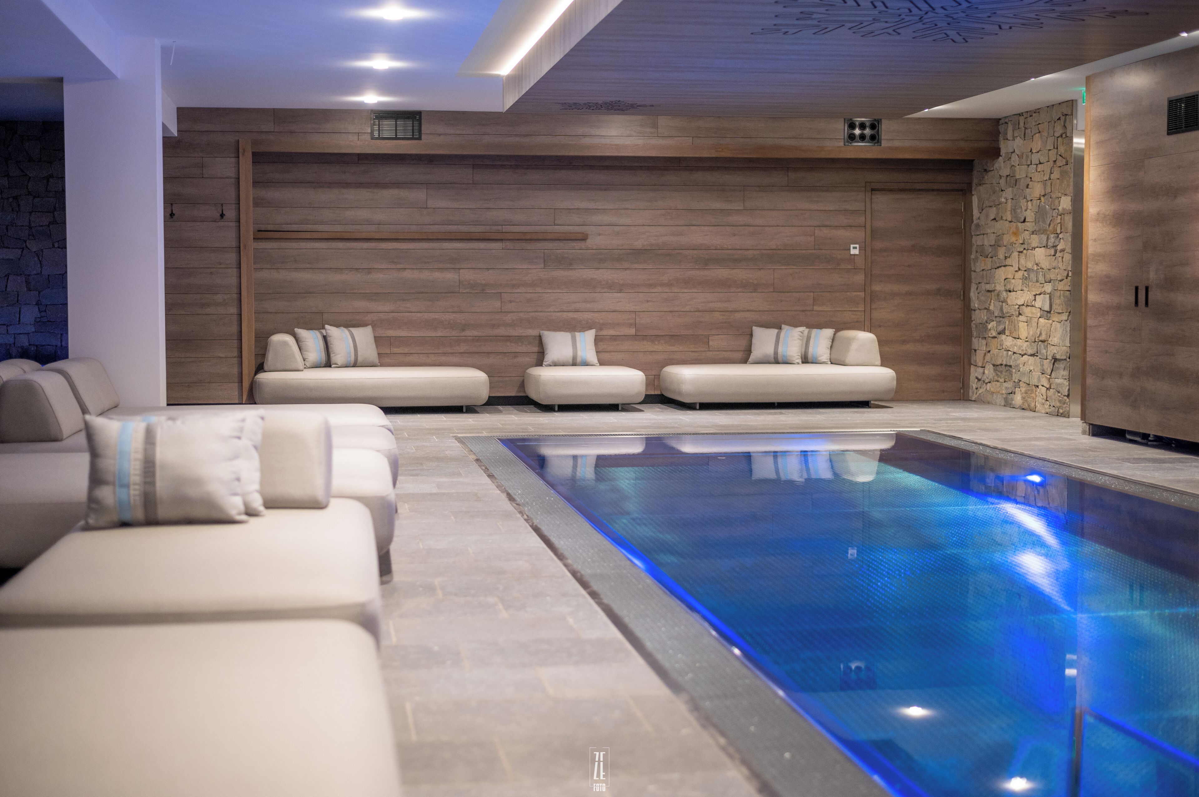 Un área interior de piscina moderna con una piscina larga y estrecha, un espacio de descanso con sofás y una pared de piedra.