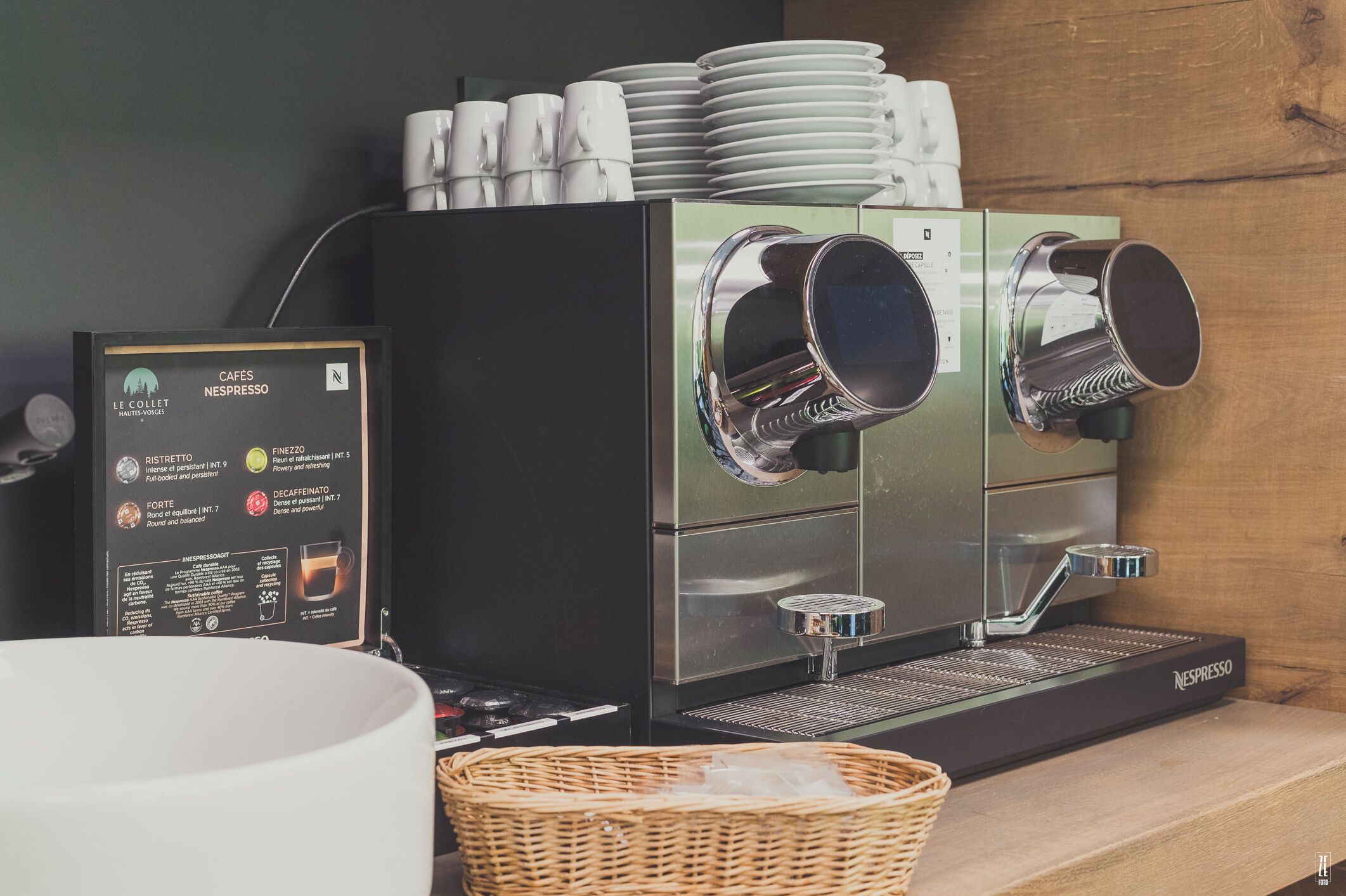Eine Nespresso-Kaffeemaschine mit mehreren Siebträgern, eine Speisekarte und ein Korbwäscher auf einer Holztresen.