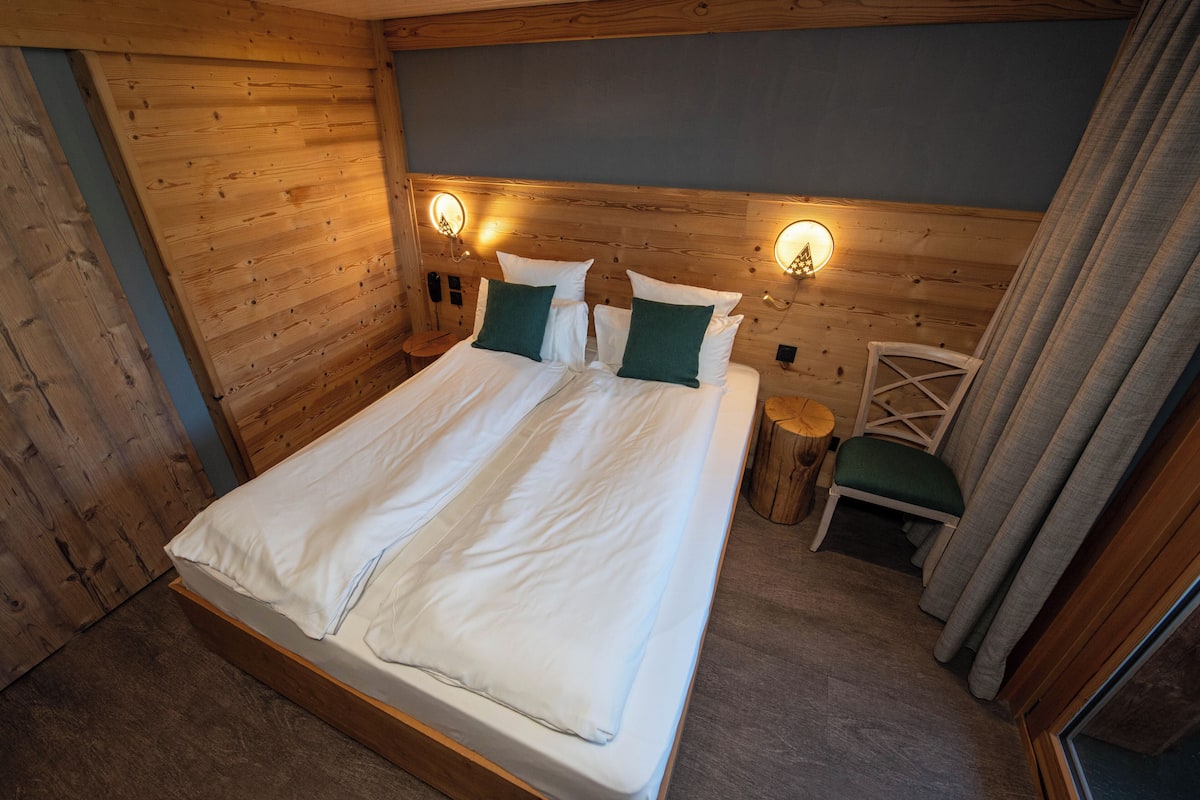 A wooden-paneled bedroom with a bed, two wall-mounted lights, a wooden chair, and a small round table.